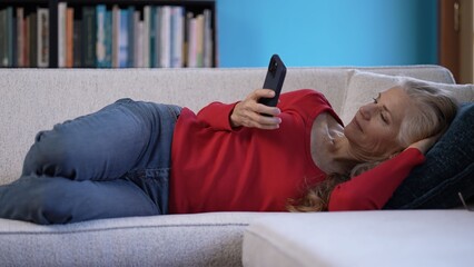 A mature woman lounges on a sofa, dressed in a red top and jeans, focused on her smartphone in a...