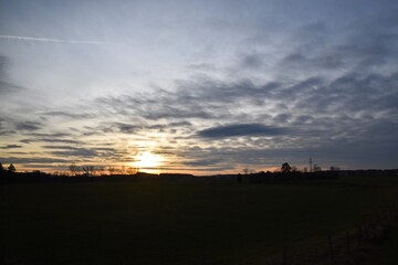 A dramatic and atmospheric sunset sky with vibrant orange and yellow clouds over a silhouetted rural landscape in Fussen, Germany. The tranquil scene captures the peaceful beauty of twilight.