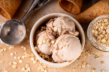 Vegan oats milk non-dairy ice cream, scoops of vanilla creamy oatmeal milk gelato in beige bowl, with ice cream waffle cones and rolled oats flakes