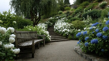 Obraz premium Gravel path winds through a serene garden past a rustic wooden bench and white hydrangeas, stone steps climbing a green hillside dotted with blue blooms for a peaceful escape