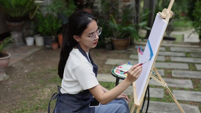 asian female artist painting outdoor in garden during leisure time holding brush sitting on chair looking at canvas with peaceful expression enjoying creative activity in backyard studio surrounded by