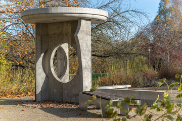 Modern concrete pavilion with circular cutouts and benches in a park. Late-autumn shrubs and bare branches surround it.
