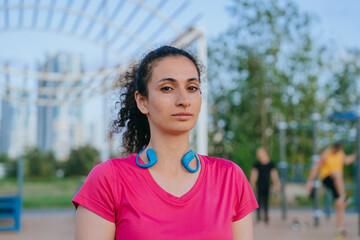 Close-up of a woman with headphones resting on her shoulders, exuding confidence in a vivid pink top against a blurred workout scene.