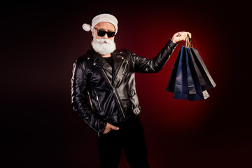 Cool Santa rocker in leather jacket with shopping bags posing against red background for festive Christmas stock photo