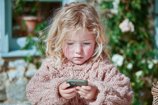 A young girl with blonde curly hair is intently focused on a smartphone in her hands outdoors