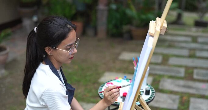 young asian artist enjoying outdoor painting session smiling slightly while holding brush adding detail to canvas in peaceful backyard surrounded by natural plants expressing creativity with calm conc
