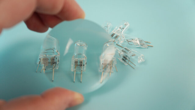 Halogen capsule lamps and a hand with a lens on a turquoise background. The magnified markings and contacts are visible through the lens, and the subject is shot from above.