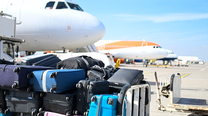 Stacked luggage handling carts with unclaimed or delayed baggage on a busy airport tarmac. Airplanes and clear skies in the background. Travel, aviation, and lost luggage themes. © tannujannu