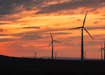 Obraz premium Multiple wind turbines silhouetted against a vibrant sunset sky. The turbines stand tall against the backdrop of orange and red clouds.