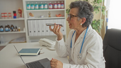Senior woman doctor with grey hair in a clinic office, wearing a stethoscope, using a smartphone to send a voice message while working on a laptop.