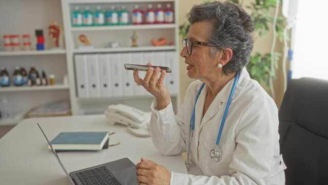 Senior woman doctor with grey hair in a clinic office, wearing a stethoscope, using a smartphone to send a voice message while working on a laptop. - Powered by Adobe