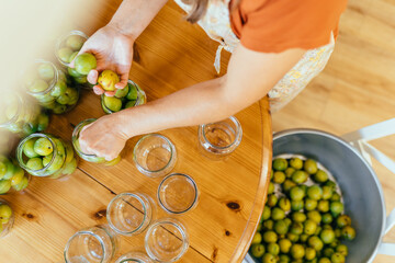 Woman placing green plums into glass jars for homemade preservation