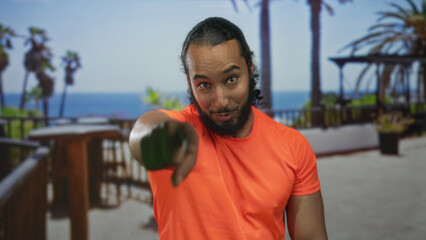 Fototapeta premium Man points finger to camera on building terrace, bare hand extended wearing orange shirt and beard; confidence invitation.