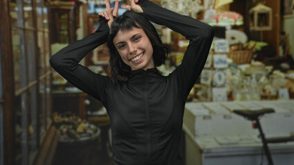 Hispanic woman shows hands making bunny ears gesture in a home decor shop filled with vintage furniture and decor items; playfulness.