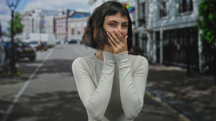 Young hispanic woman covers mouth with both hands on a sunlit bustling urban street; bashfulness...
