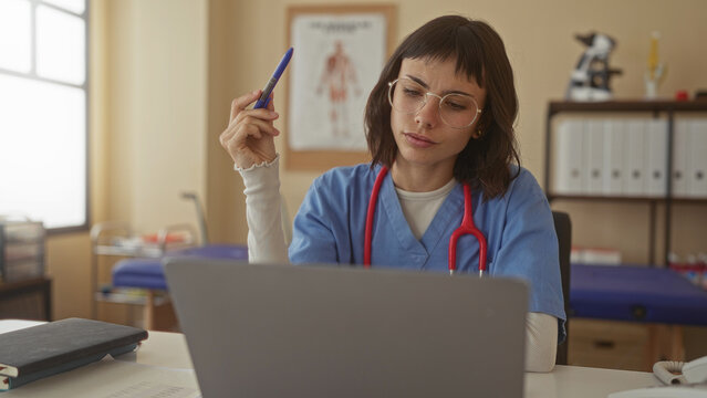 Woman holding pen at laptop in clinic with red stethoscope hanging on neck and anatomical chart behind; concentration.