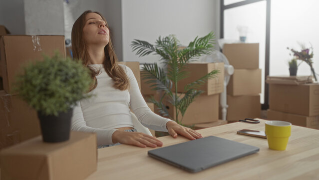 Young woman typing with hands on laptop at a wooden table surrounded by moving boxes and a potted plant in a building; moving day fatigue.