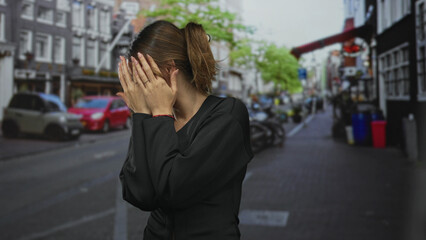 Fototapeta premium Young hispanic woman clasping hands to chest on a busy city street with parked cars and bicycles visible; fear caution.