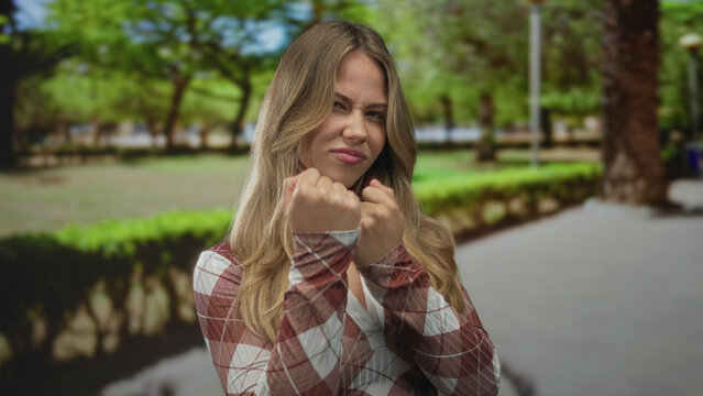 Woman thrusts bare fist forward in green sunlit park with swaying blonde hair; confidence determination empowerment. - Powered by Adobe