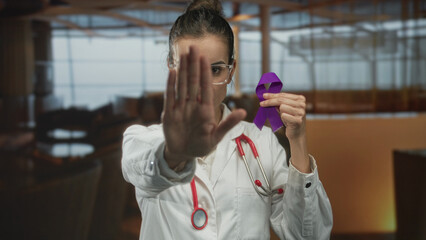 Woman doctor with stethoscope holding purple ribbon and palm forward stop gesture in building; solidarity determination.