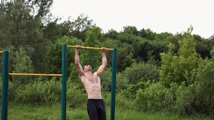 Muscular athlete doing pull ups on horizontal bar at sports ground. Young shirtless sportsman exercising at nature. Strong sporty man training outdoor. Concept of healthy and active lifestyle. Slow mo