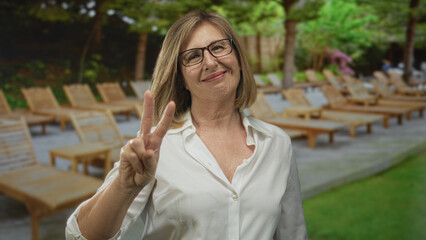 Fototapeta premium Woman wearing glasses and white shirt makes peace sign in front of a building with lounge chairs and greenery, leaning forward with a gentle smile; serenity.