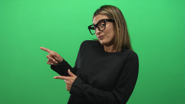 Young blonde woman in studio pointing index finger to the right, wearing black sweater and oversized glasses against green background; skeptical attitude. - Powered by Adobe