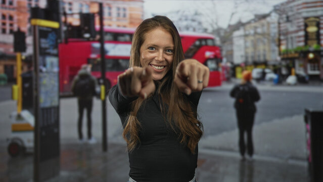 Woman points fingers at camera on bustling city street with red doubledecker bus and pedestrians in view; confidence invitation connection.