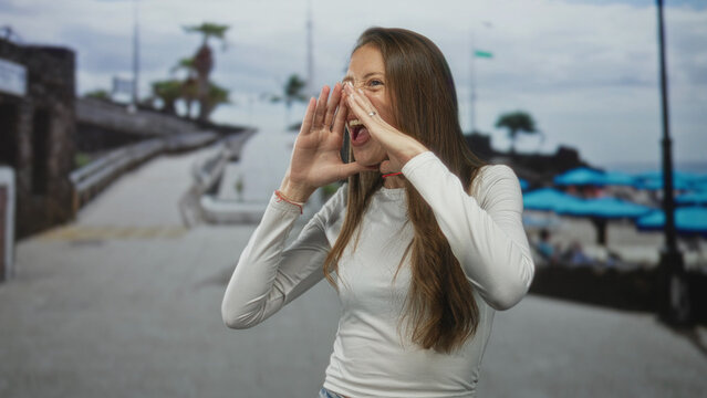 Woman cupping hands to mouth calling out along a coastal beach promenade beside blue umbrellas and a stone boardwalk; joy wanderlust spontaneity.