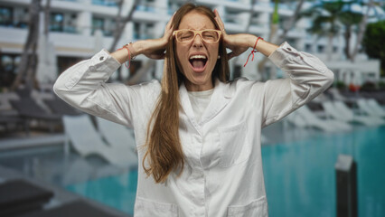Young hispanic woman in labcoat holding hands to head screaming at hotel pool; overwhelmed...