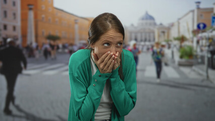 Fototapeta premium Woman hands covering mouth on a crowded street with cathedral and crosswalk visible, forehead wrinkles and rings on fingers; surprise.