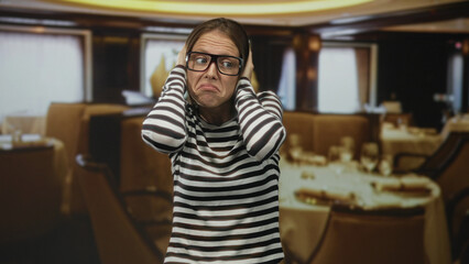 Woman covers ears with hands in building restaurant interior wearing striped shirt and glasses;...