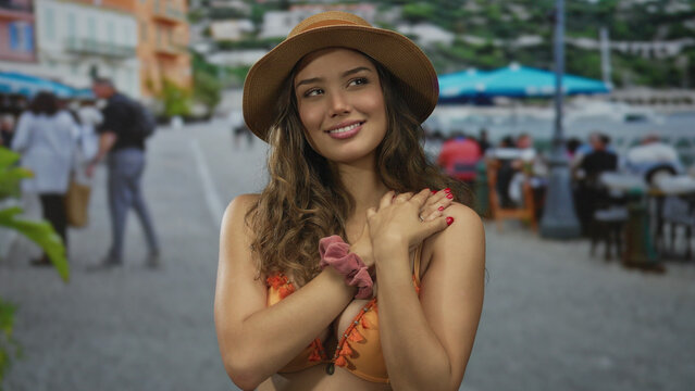 Young hispanic woman in sunhat and orange bikini makes finger heart gesture on street outside seaside restaurant; affection. - Powered by Adobe