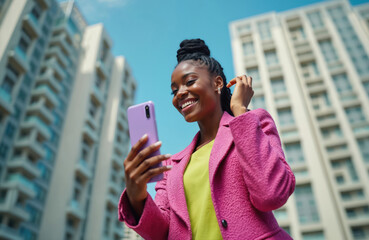 Young black woman holds purple smartphone, takes selfie with bright smile. She wears pink jacket outdoors near tall modern buildings. Blue sky above, cheerful mood.