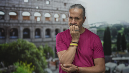 Man biting nails with hand to mouth at the roman coliseum building outdoors, long hair and...
