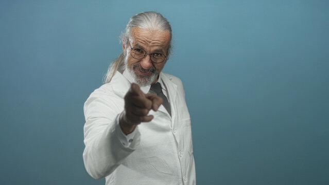 Scientist man pointing finger in blue studio setting, smiling in white lab coat and glasses; confidence expertise scientific authority.