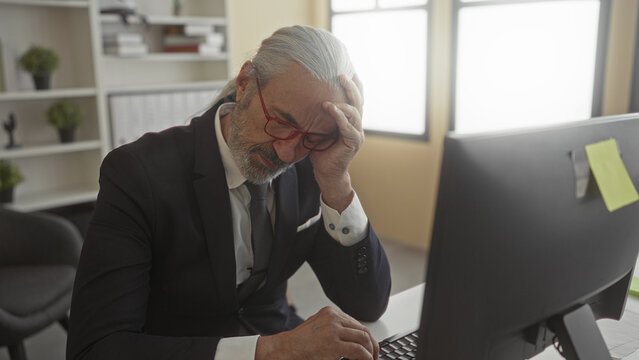 Man in suit typing at computer with hand on forehead for headache in office building; stress fatigue. - Powered by Adobe