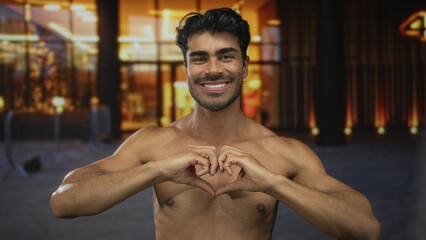 Fototapeta premium Young hispanic shirtless man forming heart shape with hands under street lights on busy urban street at dusk; love.