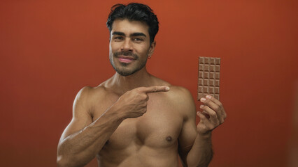 Fototapeta premium Muscular shirtless man points finger to a chocolate bar while smiling in an orange studio; temptation indulgence.
