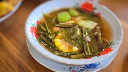 Mixed vegetable tamarind called sayur asem featuring leafy greens, corn, and potato pieces in a rich broth. Cozy, wholesome meal set on a wooden table