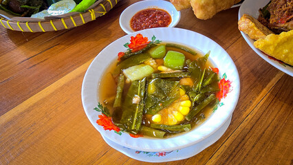 Mixed vegetable tamarind called sayur asem featuring leafy greens, corn, and potato pieces in a rich broth. Cozy, wholesome meal set on a wooden table