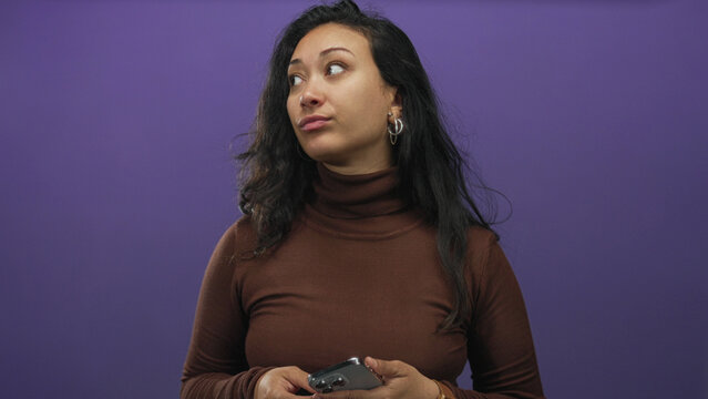 Young hispanic woman holding smartphone with both hands in purple studio while glancing at the screen; skepticism.