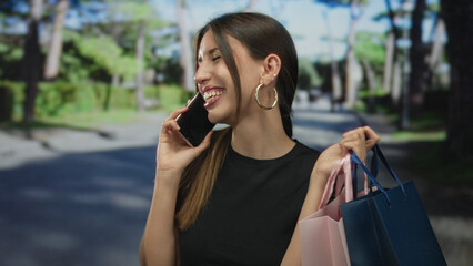 Woman holding smartphone to ear on street while carrying pastel shopping bags beneath leafy green trees; happiness.