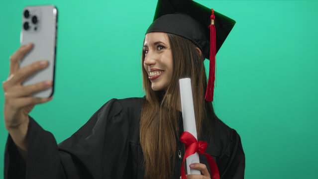 Woman in graduation gown holds smartphone for selfie with diploma and red tassel against green backdrop in studio; pride.