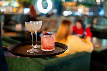 Waiter serving two vibrant cocktails on a dark tray in a stylish, dimly lit bar with blurred patrons in the background, offering a glimpse into nightlife and service