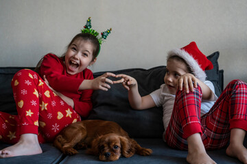 Sisters in Christmas pajamas sit on the sofa with an English King Charles puppy.