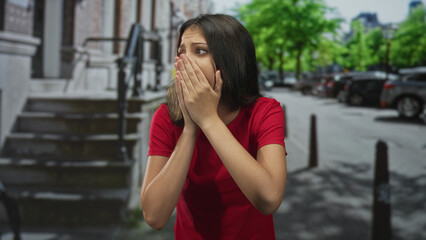 Fototapeta premium Woman covers mouth with both hands on city street, palms raised and shoulders shrugged in expressive plea; anxiety.