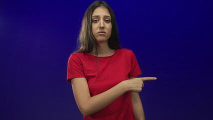Obraz premium Young woman finger pointing left in blue studio wearing red t shirt, visible hand pointing to left side; skepticism.