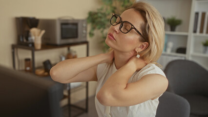 Blonde woman with glasses and eyes closed holds neck near computer in office building; stress...