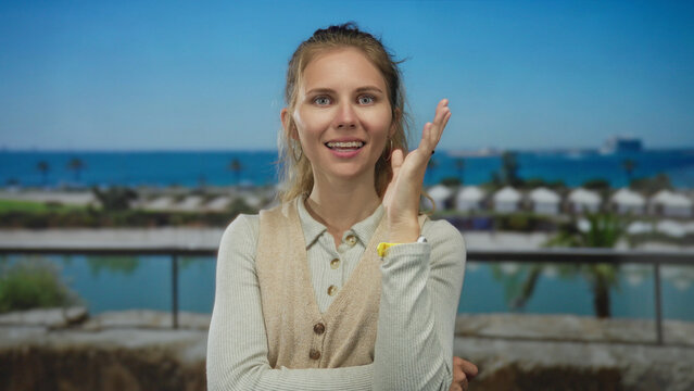 Woman rests hand on chin thinking on balcony with blonde young hair blown by wind overlooking ocean under blue scenery wearing casual clothing.